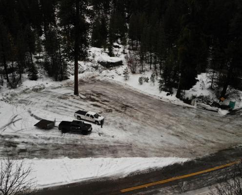 Plowed parking area with two trucks in the middle, next to a plowed highway surrounded by pine trees. A porta-potty stands in the corner.